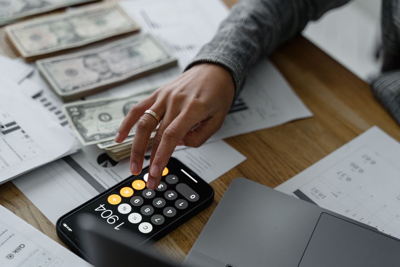 Close-up of a person using a smartphone calculator amid money and financial documents on a wooden table.